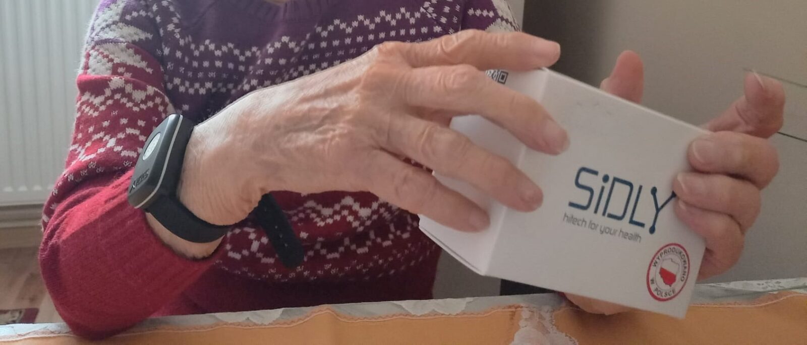 20220405 opaski 2022: Na zdjęciu widać seniora, który ma na ręce opaskę telemedyczną, a w dłoniach trzyma opakowanie od urządzenia producenta opaski telemedycznej firmy SIDLY