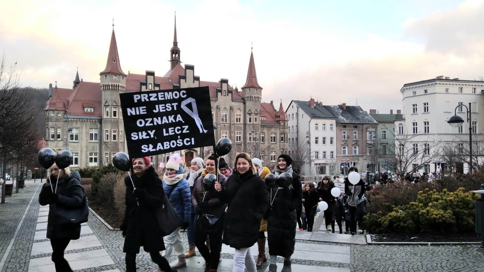 [Tekst ALT: zdjęcie przedstawia plac Magistracki i Ratusz, na tle którego widoczna jest grupa osób biorąca udział w marszu, trzymająca plakat z hasłem: przemoc nie jest oznaką siły lecz słabości. Obok hasła namalowany jest symbol białej wstążki]